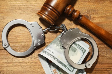 Handcuffs with dollar banknotes and gavel on wooden table, flat lay