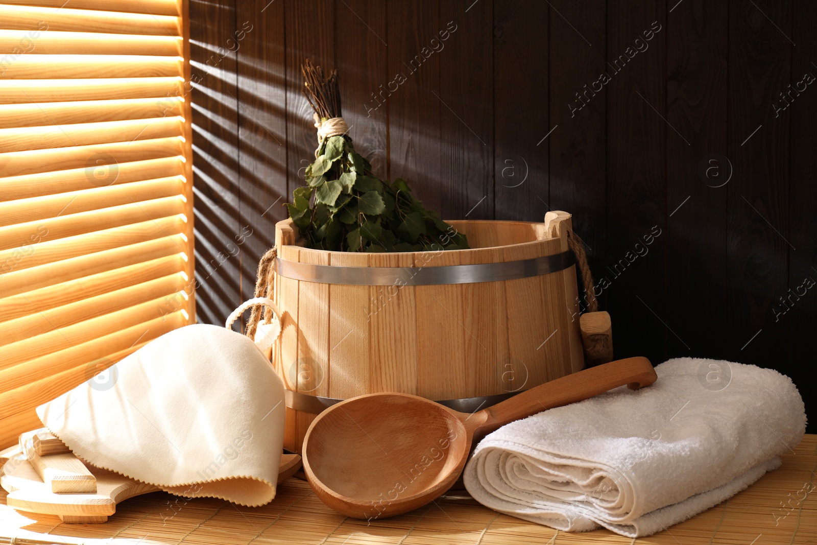 Photo of Sauna equipment. Bucket, ladle, felt wool hat, birch whisk and towels on bamboo bench indoors