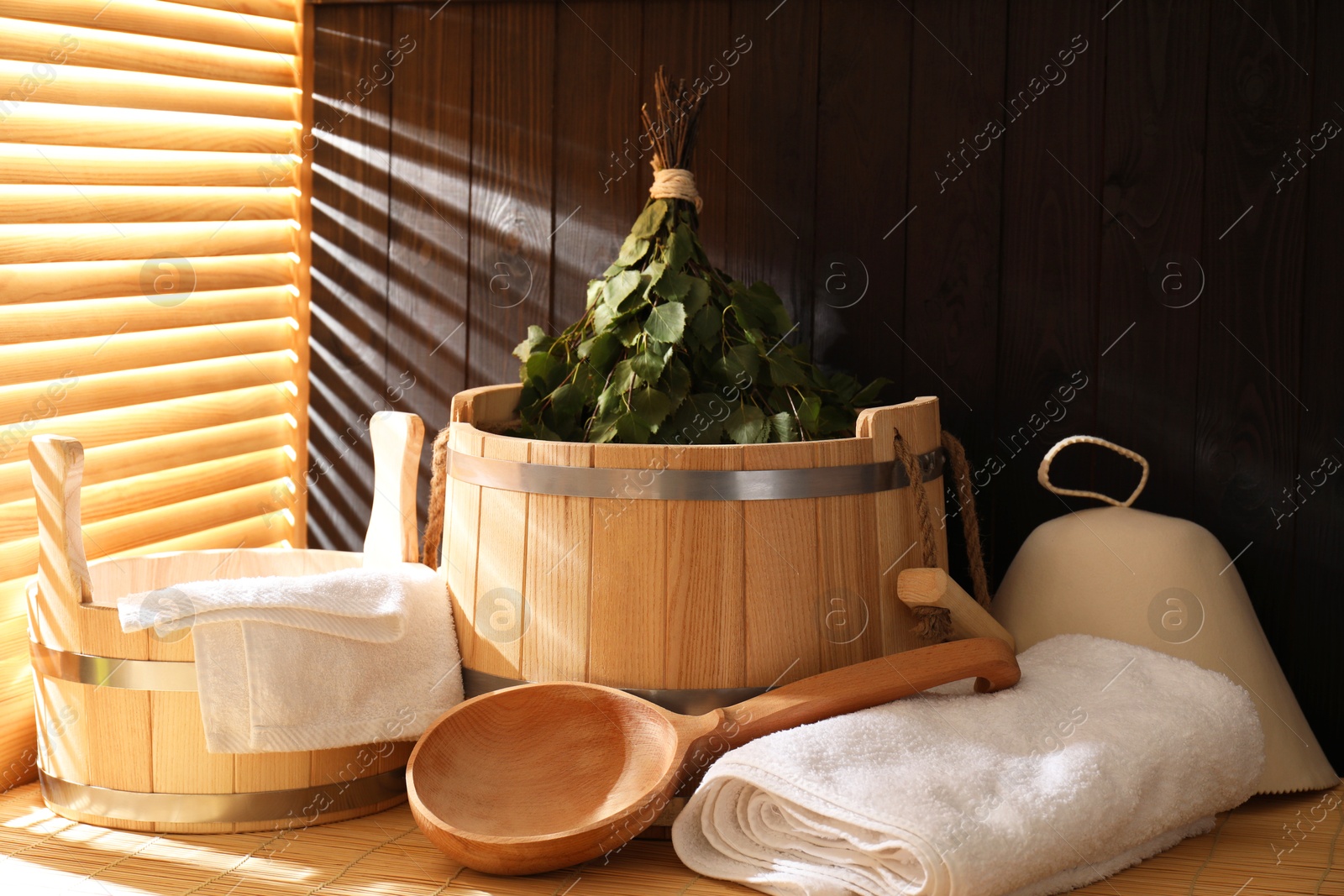 Photo of Sauna equipment. Bucket, ladle, felt wool hat, birch whisk and towels on bamboo bench indoors