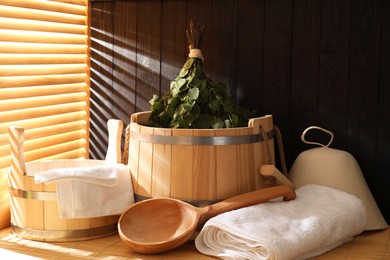 Photo of Sauna equipment. Bucket, ladle, felt wool hat, birch whisk and towels on bamboo bench indoors