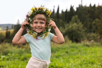 Photo of Portrait of smiling little girl with floral wreath at meadow, space for text. Child enjoying beautiful nature