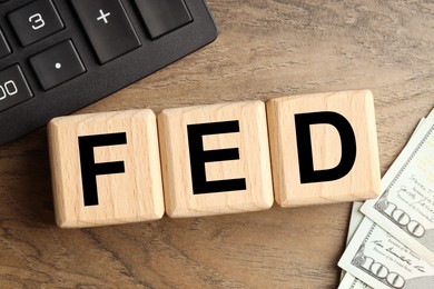 Photo of Cubes with letters Fed (Federal Reserve System), dollar banknotes and calculator on wooden table, flat lay