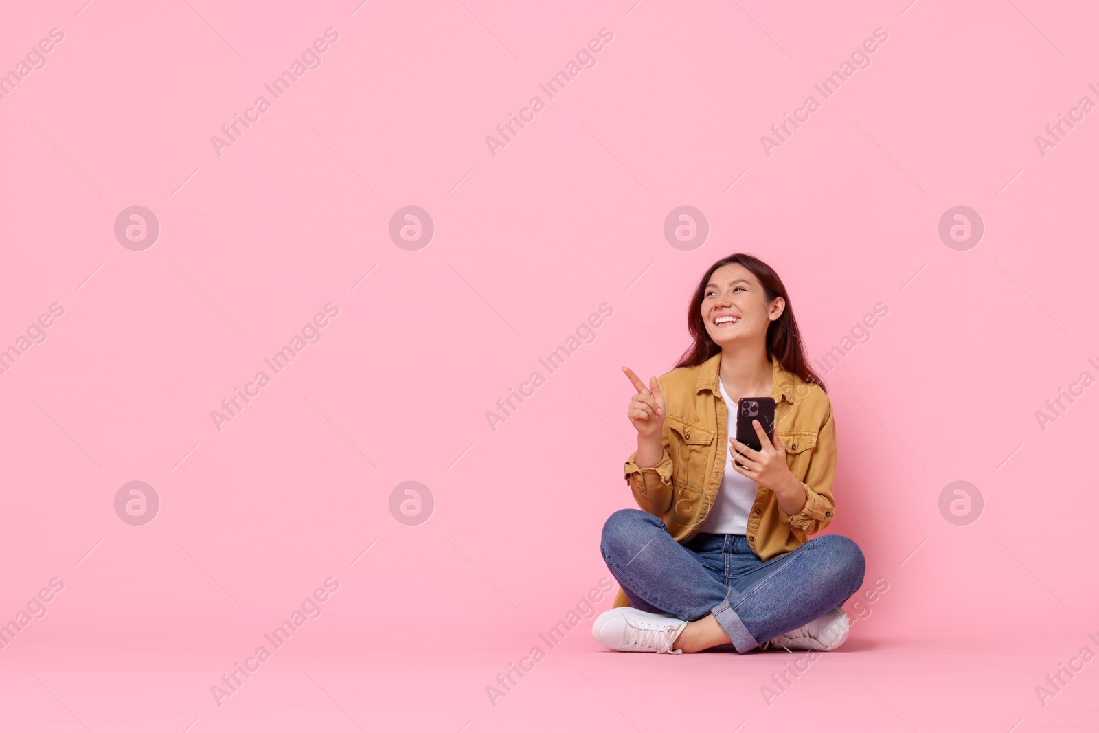 Photo of Smiling young woman with smartphone pointing at something on pink background. Space for text