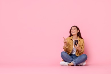 Photo of Smiling young woman with smartphone pointing at something on pink background. Space for text