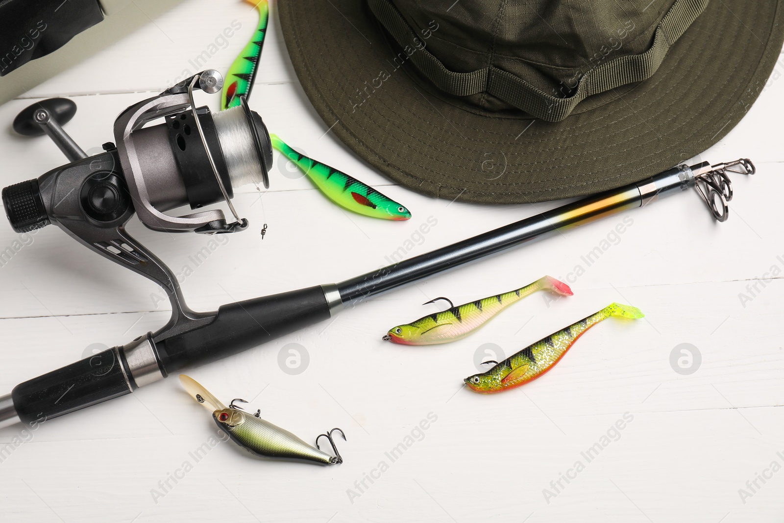 Photo of Fishing rod with spinning reel, hat and baits on white wooden table, flat lay