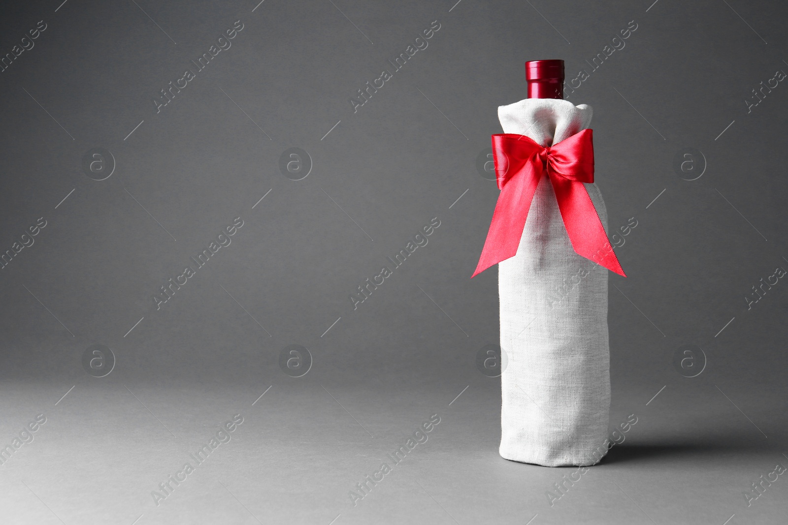 Photo of Wine bottle in burlap package with red ribbon on grey background, space for text