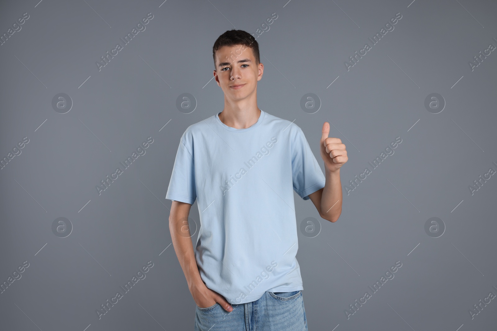 Photo of Teenage boy wearing light blue t-shirt and showing thumbs up on grey background