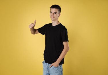 Photo of Teenage boy wearing black t-shirt and showing thumbs up on yellow background