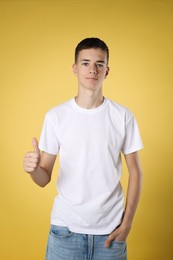 Photo of Teenage boy wearing white t-shirt and showing thumbs up on yellow background