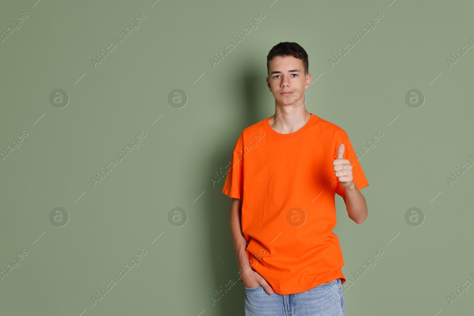 Photo of Teenage boy wearing orange t-shirt and showing thumbs up on green background, space for text