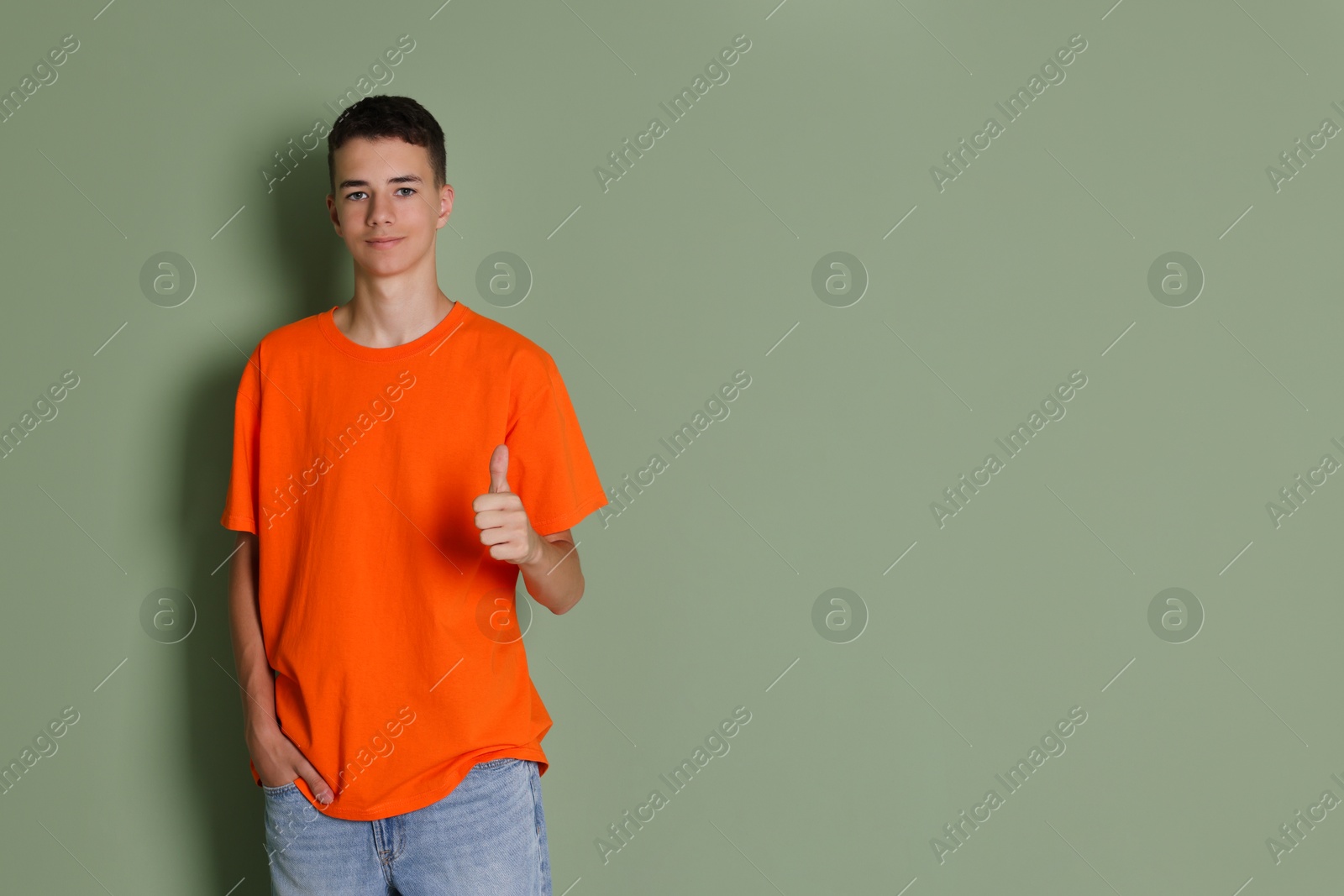 Photo of Teenage boy wearing orange t-shirt and showing thumbs up on green background, space for text