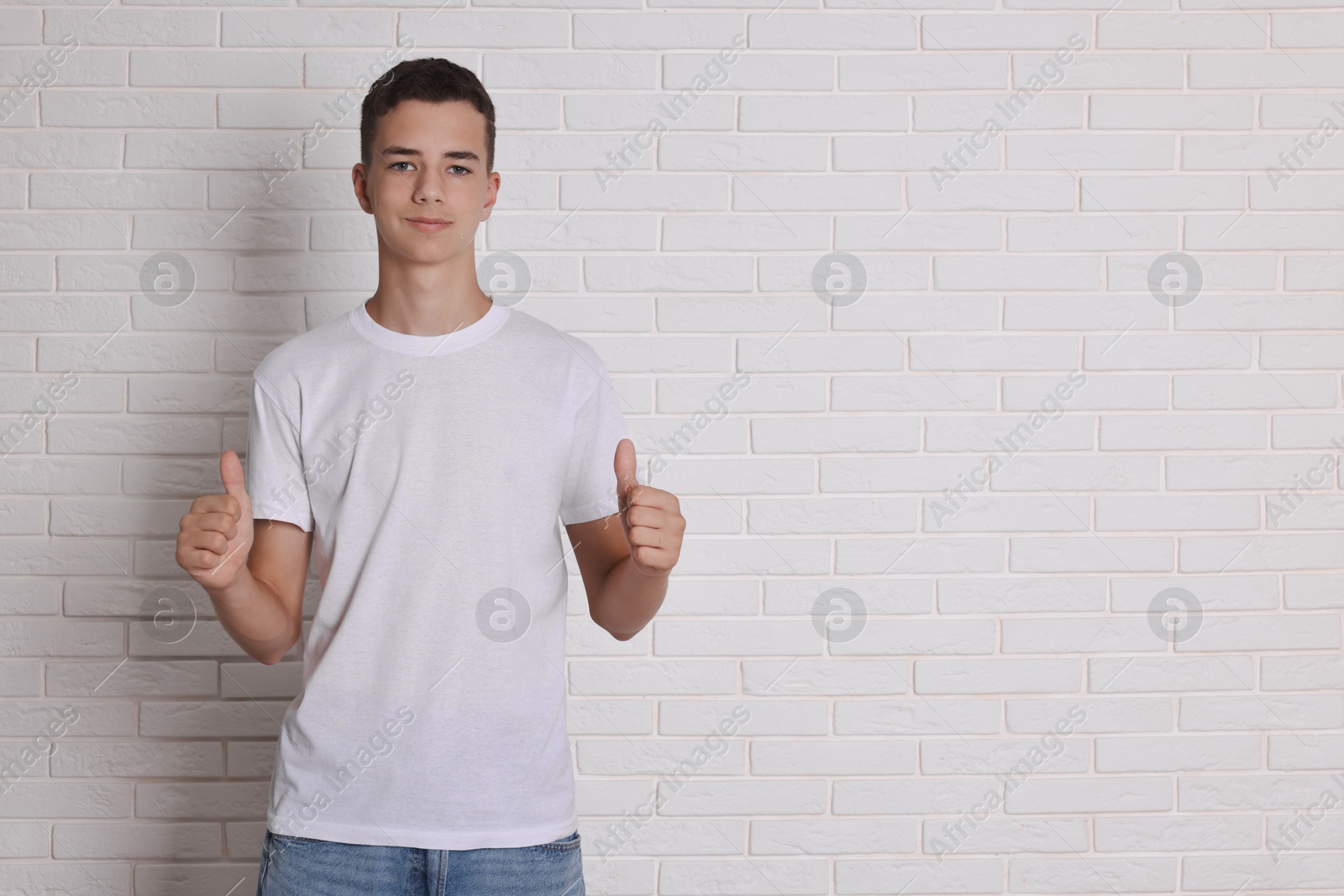 Photo of Teenage boy wearing t-shirt and showing thumbs up near white brick wall, space for text