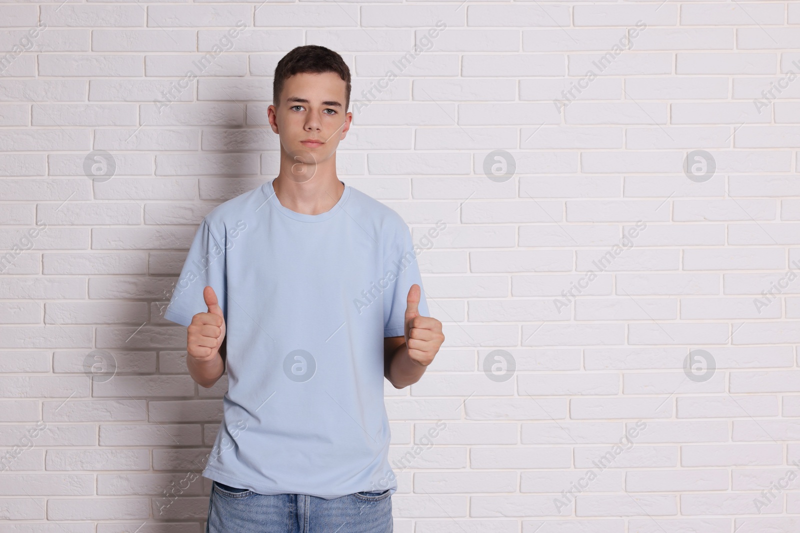 Photo of Teenage boy wearing light blue t-shirt and showing thumbs up near white brick wall, space for text