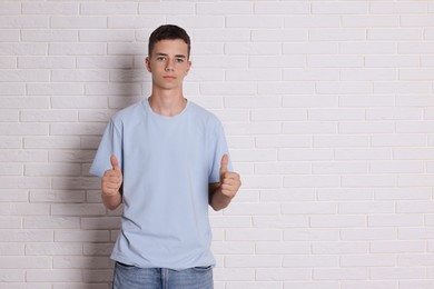 Photo of Teenage boy wearing light blue t-shirt and showing thumbs up near white brick wall, space for text