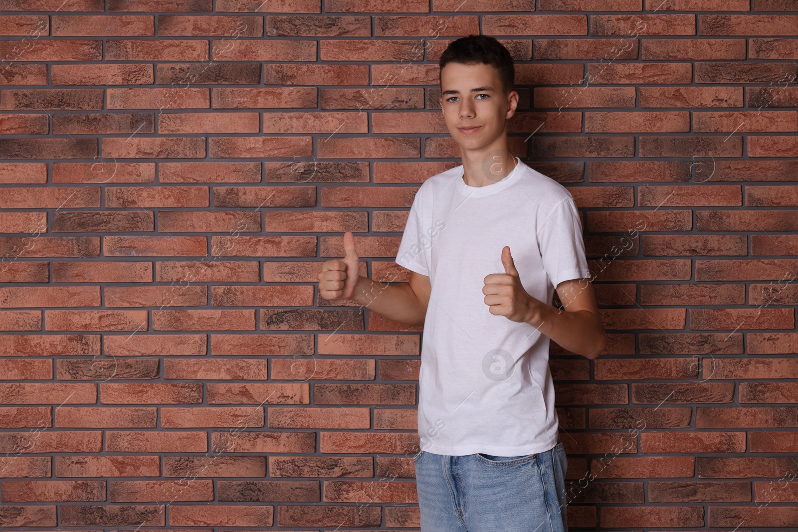 Photo of Teenage boy wearing white t-shirt and showing thumbs up near brick wall, space for text