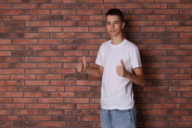 Photo of Teenage boy wearing white t-shirt and showing thumbs up near brick wall, space for text