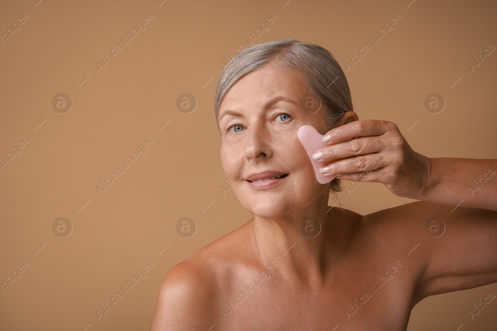 Photo of Beautiful woman doing facial massage with gua sha tool on beige background, space for text