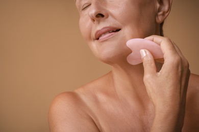 Photo of Woman doing facial massage with gua sha tool on beige background, closeup