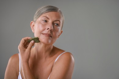 Photo of Beautiful woman doing facial massage with gua sha tool on grey background, space for text