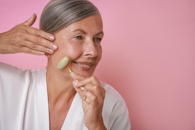 Beautiful woman doing facial massage with roller on pink background, space for text