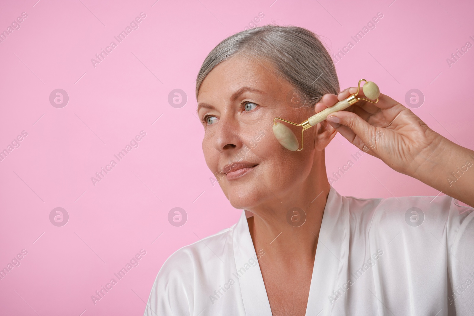 Photo of Beautiful woman doing facial massage with roller on pink background