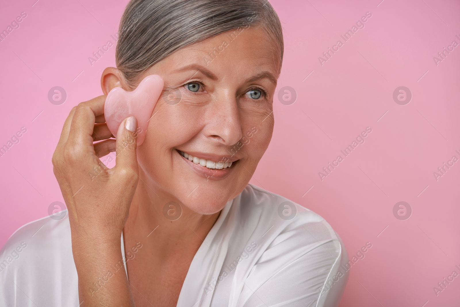 Photo of Beautiful young woman doing facial massage with gua sha tool on pink background