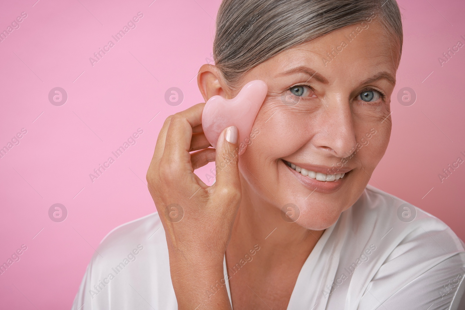 Photo of Beautiful young woman doing facial massage with gua sha tool on pink background