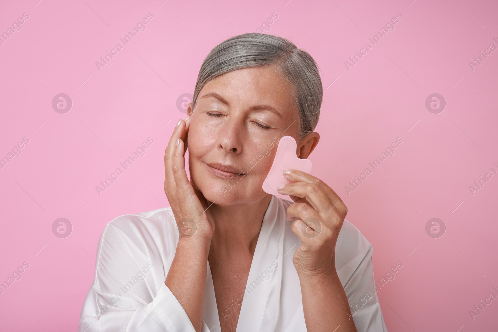 Photo of Beautiful young woman doing facial massage with gua sha tool on pink background