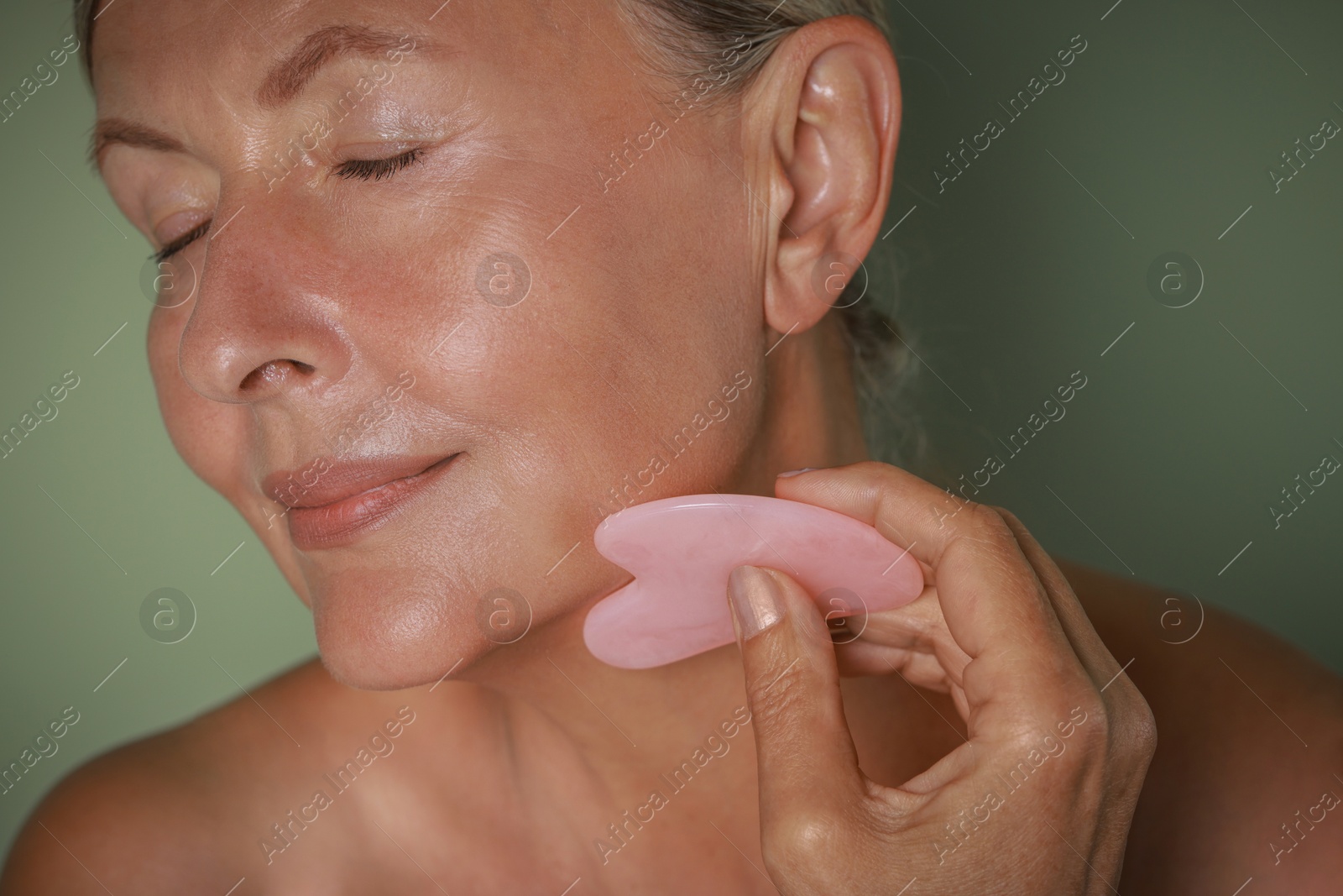 Photo of Beautiful young woman doing facial massage with gua sha tool on light green background, closeup