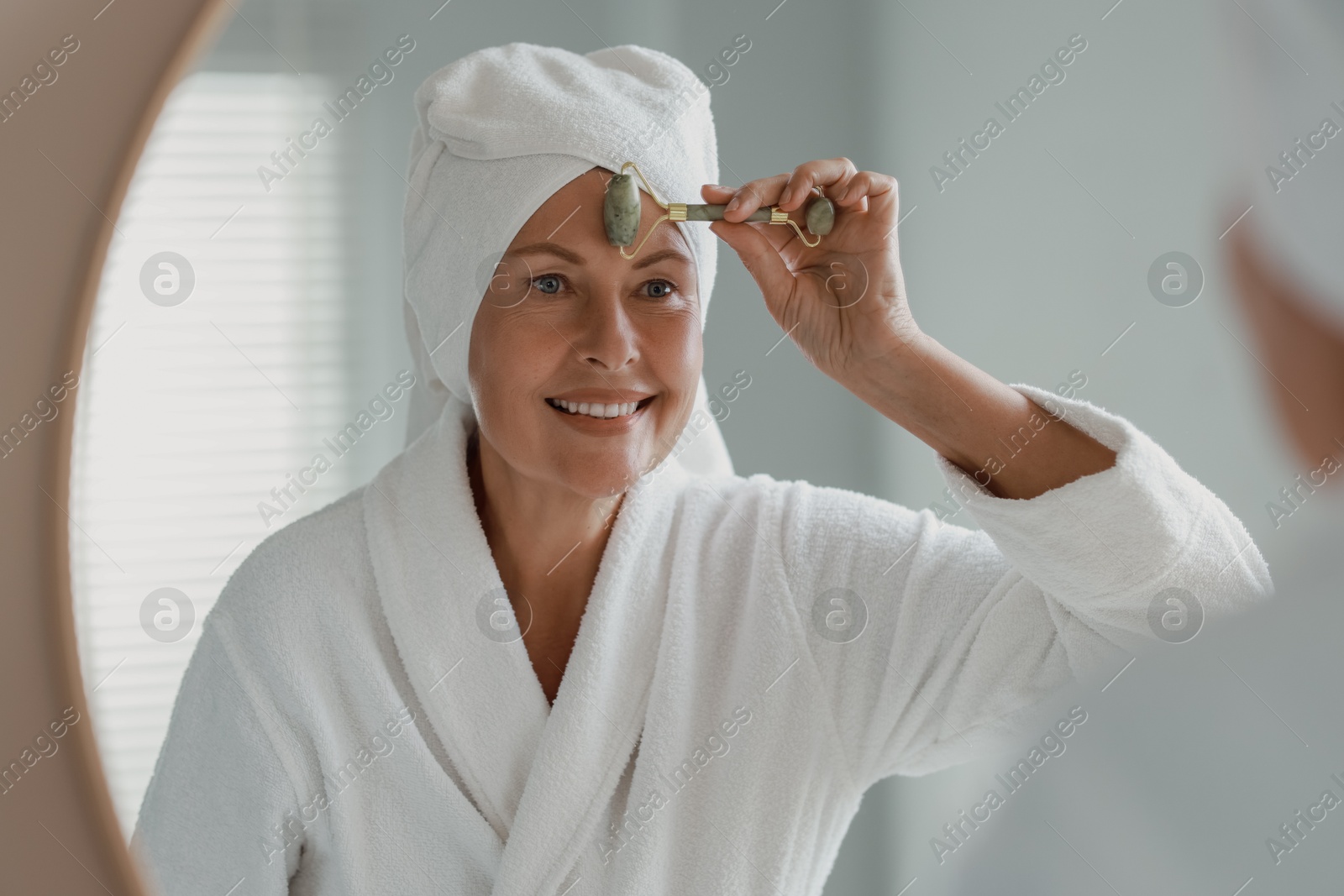 Photo of Beautiful woman doing facial massage with roller near mirror at home