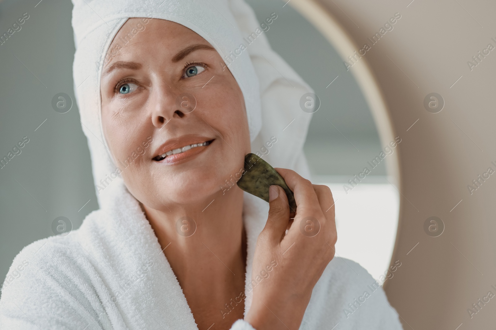 Photo of Beautiful young woman doing facial massage with gua sha tool indoors