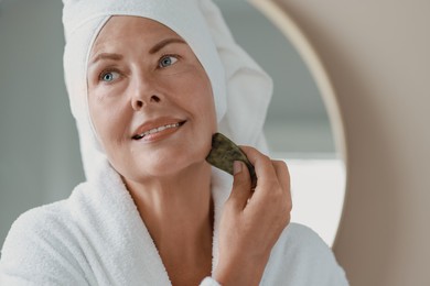 Photo of Beautiful young woman doing facial massage with gua sha tool indoors