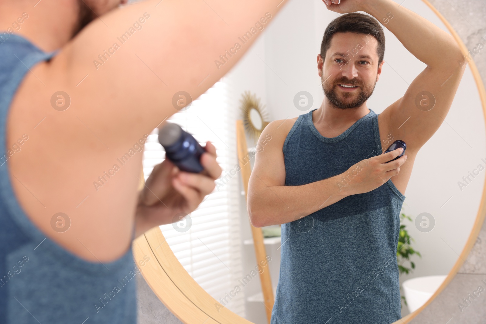 Photo of Smiling man applying roll-on deodorant near mirror at home