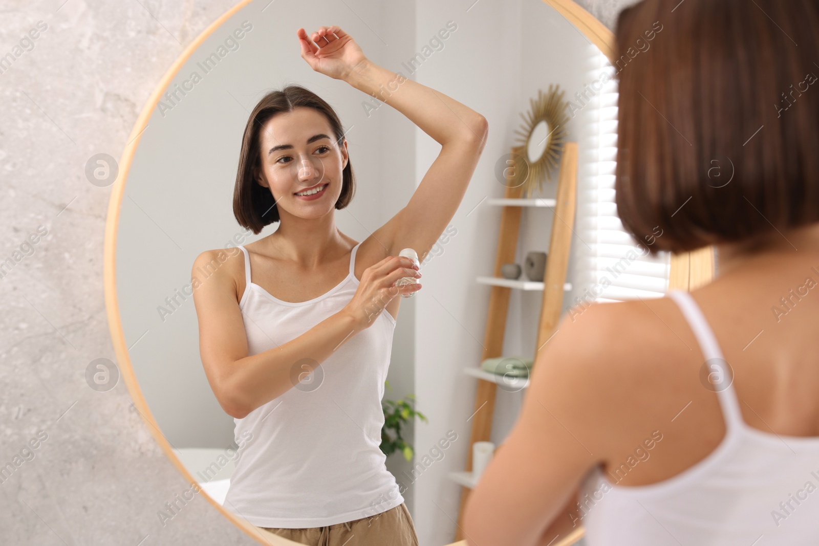 Photo of Smiling woman applying roll-on deodorant near mirror at home