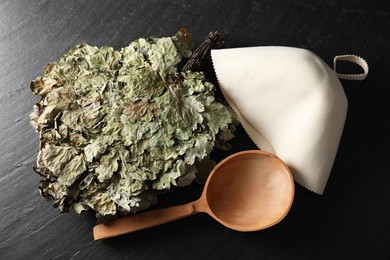 Photo of Sauna equipment. Oak whisk, felt wool hat and wooden ladle on black textured surface, flat lay