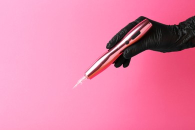 Photo of Professional cosmetologist holding permanent makeup machine on pink background, closeup. Space for text