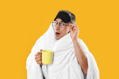 Photo of Overslept man with sleep mask, glasses and cup wrapped in blanket on orange background