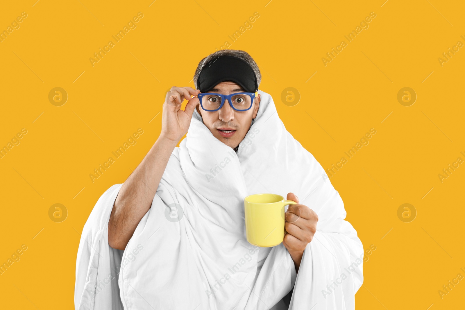 Photo of Overslept man with sleep mask, glasses and cup wrapped in blanket on orange background