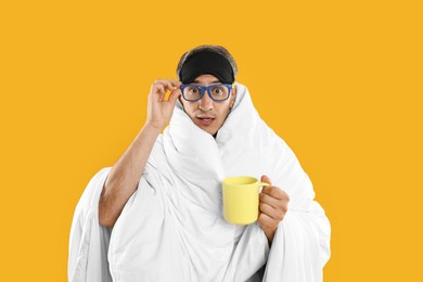 Photo of Overslept man with sleep mask, glasses and cup wrapped in blanket on orange background