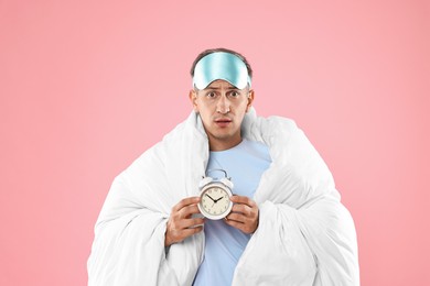 Overslept man with sleep mask and alarm clock wrapped in blanket on pink background