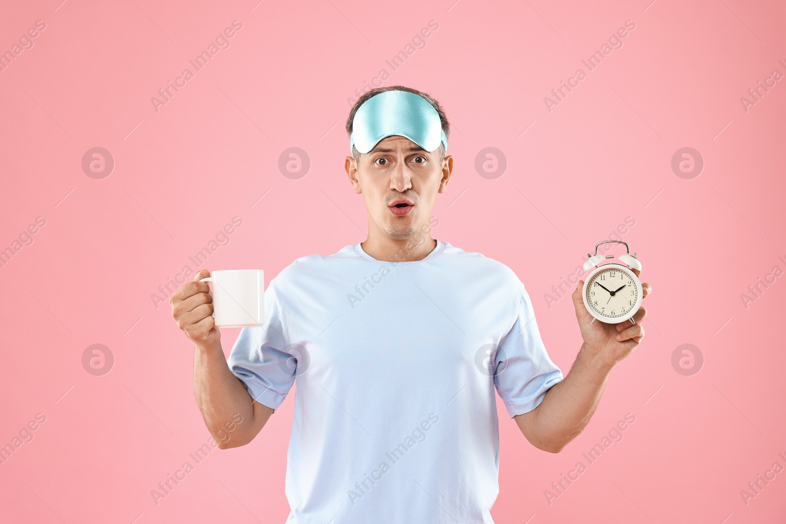 Photo of Overslept man with sleep mask, cup and alarm clock on pink background