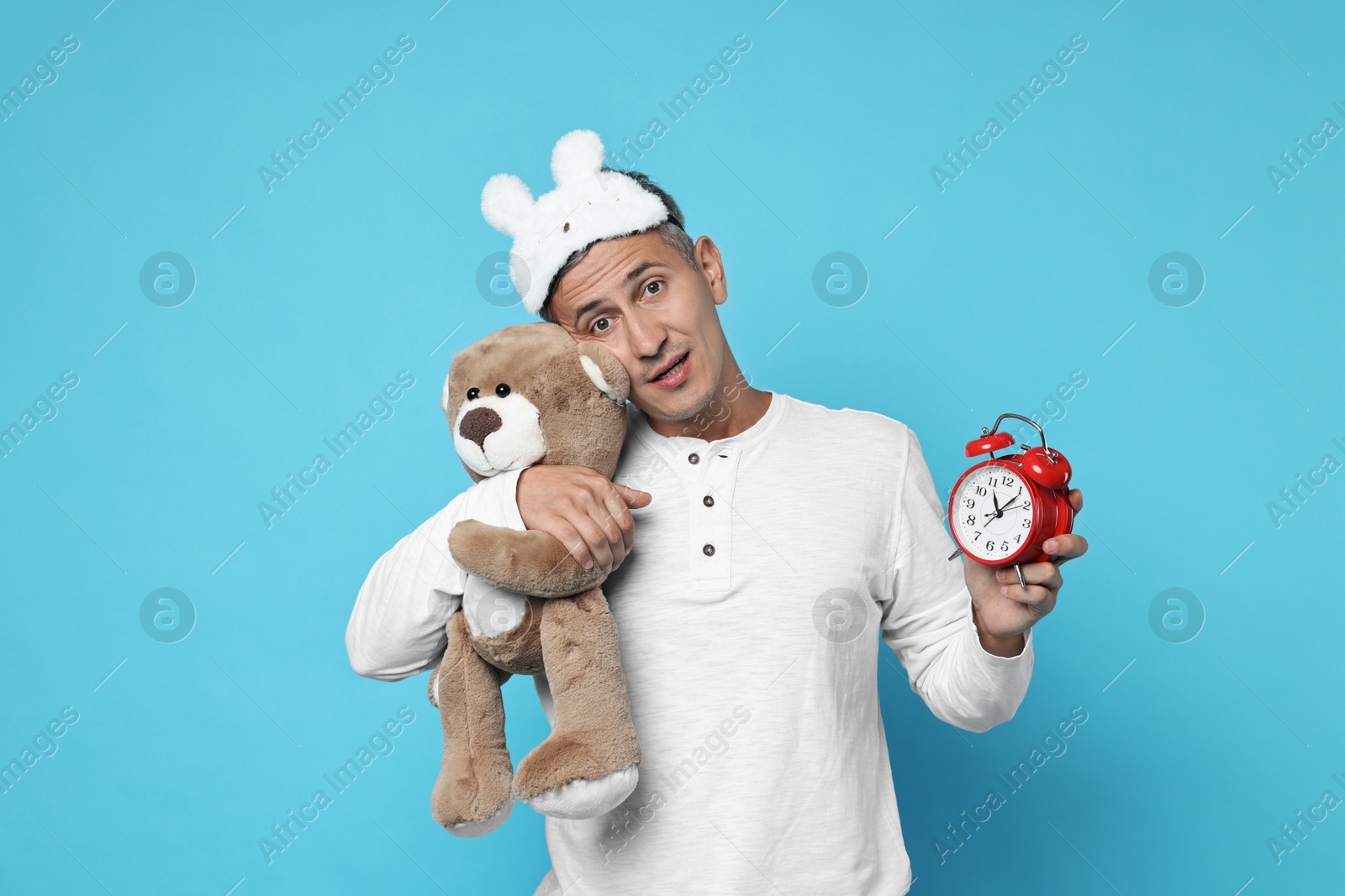 Photo of Overslept man with sleep mask, teddy bear and alarm clock on light blue background