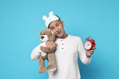 Photo of Overslept man with sleep mask, teddy bear and alarm clock on light blue background