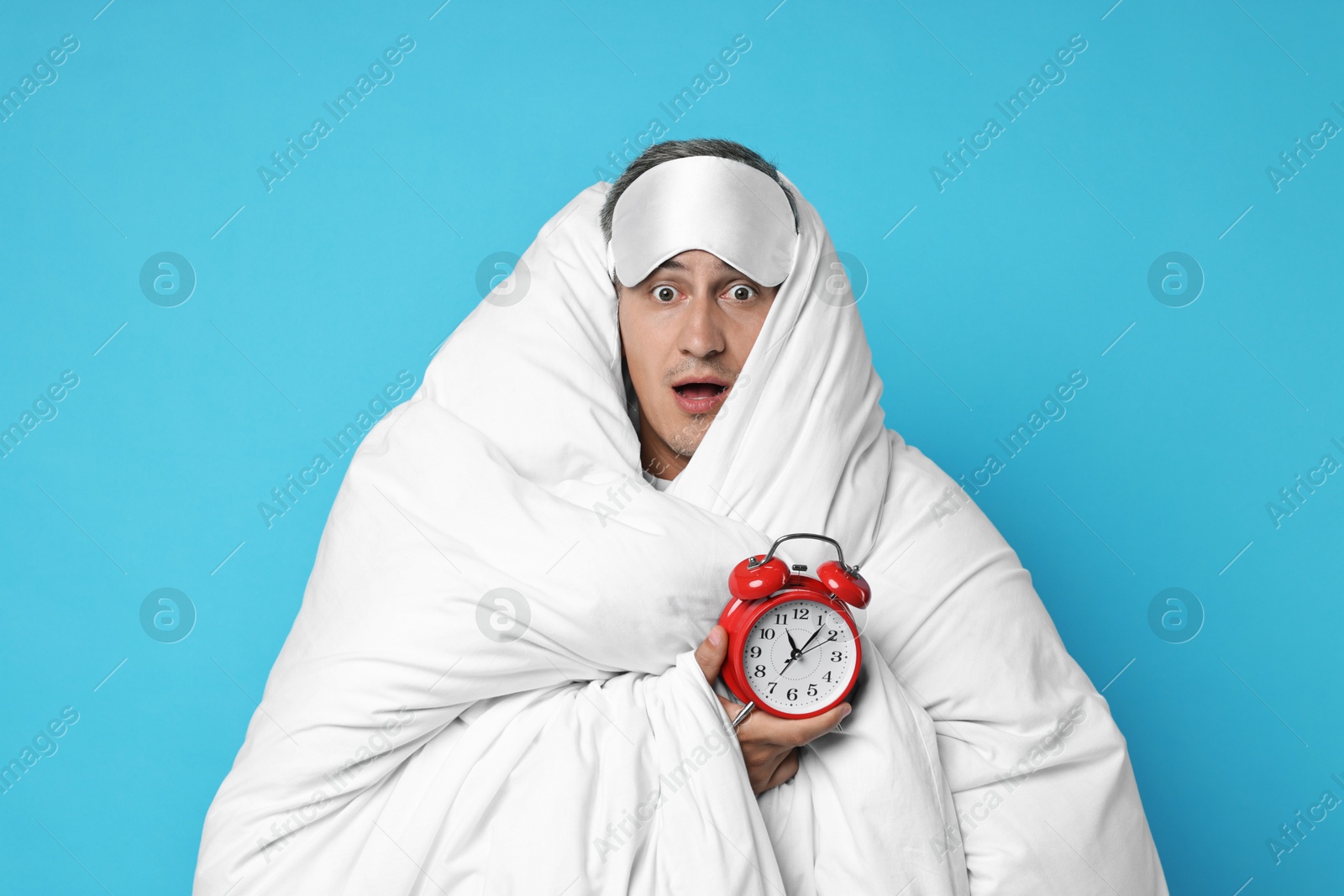 Photo of Overslept man with sleep mask and alarm clock wrapped in blanket on light blue background