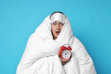 Overslept man with sleep mask and alarm clock wrapped in blanket on light blue background