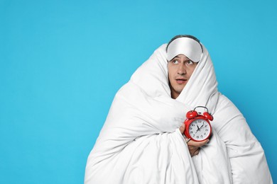 Photo of Overslept man with sleep mask and alarm clock wrapped in blanket on light blue background, space for text