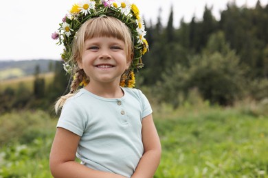 Photo of Portrait of smiling little girl in floral wreath outdoors, space for text. Child enjoying beautiful nature