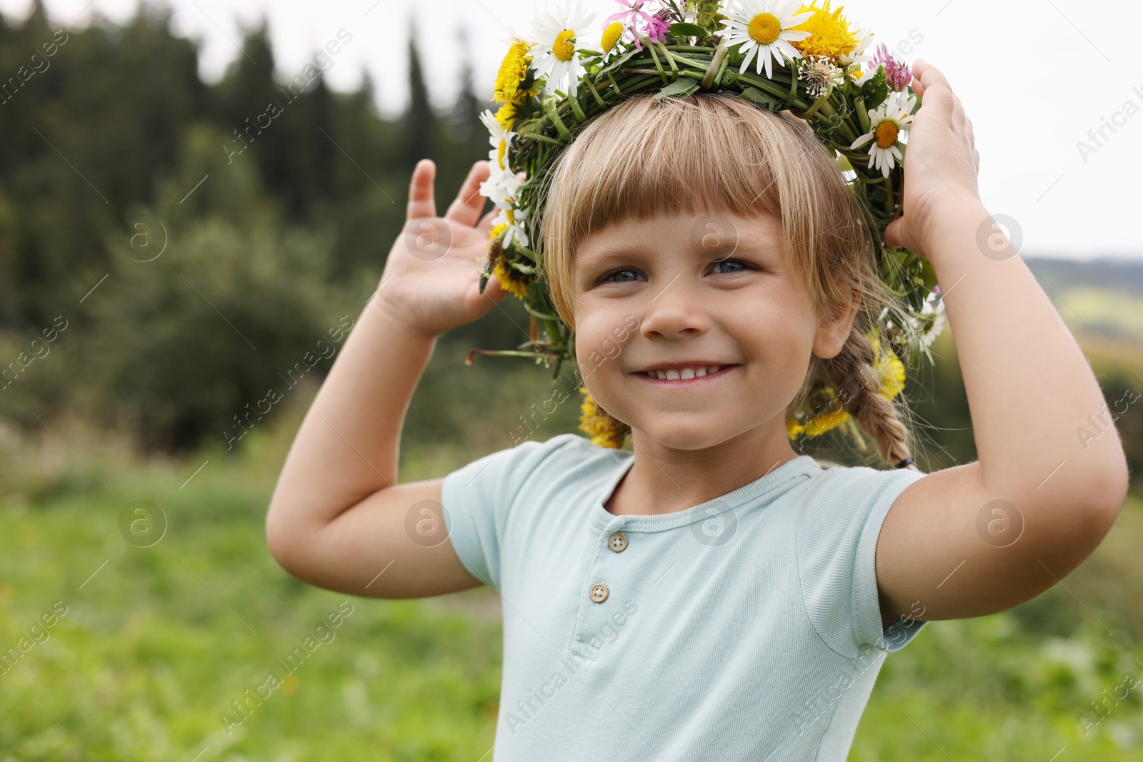 Photo of Portrait of smiling little girl in floral wreath outdoors, space for text. Child enjoying beautiful nature