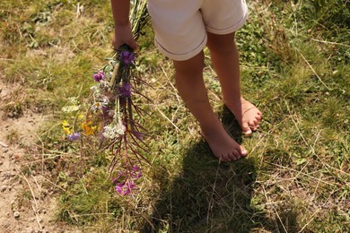 Photo of Little girl with bouquet of wildflowers walking barefoot outdoors on sunny day, closeup. Child enjoying beautiful nature