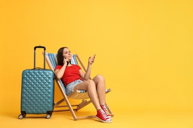 Photo of Happy young woman talking on phone and suitcase against orange background, space for text